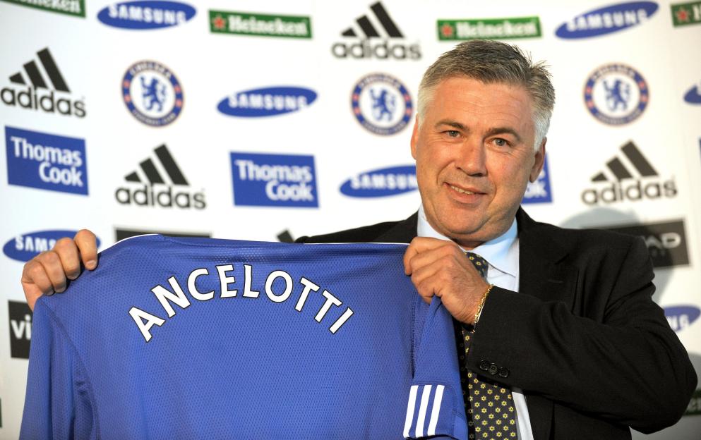 Carlo Ancelotti holding up a blue Chelsea FC jersey with "ANCELOTTI" on the back.