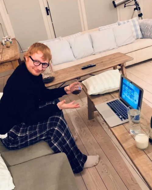 Ed Sheeran sitting on a couch in plaid pajamas, holding a glass, with a laptop and drinks on a coffee table.