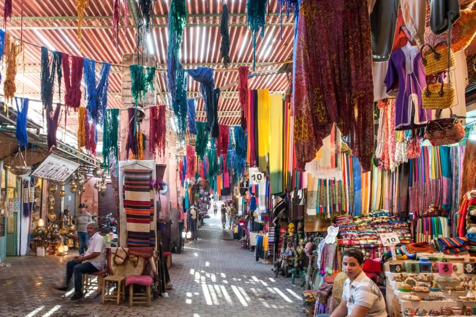 Souk in Marrakech, Morocco with colorful hanging textiles and people.