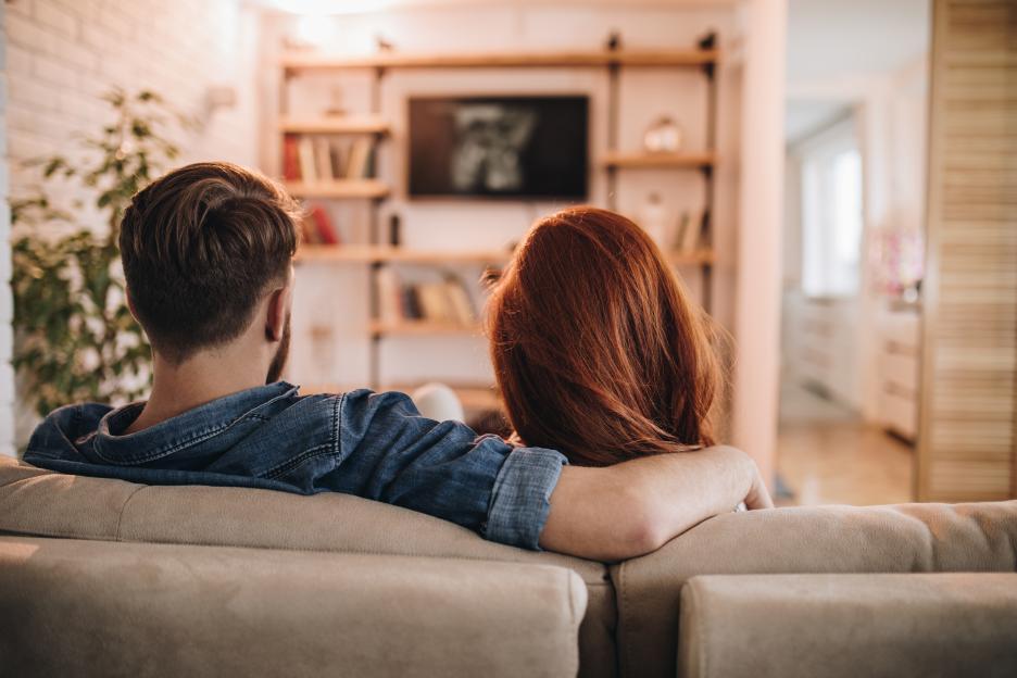 Rear view of a couple watching TV, with the man's arm around the woman.