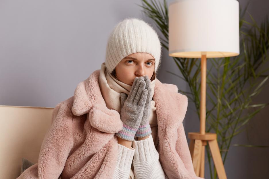 A woman in a winter coat, hat, and mittens warms her hands in a cold house.