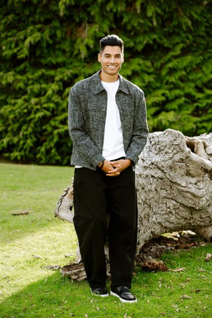 Siva Kaneswaran smiling, standing outdoors with trees and a log in the background.