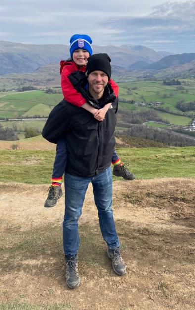 Andy Smith carries his daughter Emily on his back with a scenic valley and mountains in the background.
