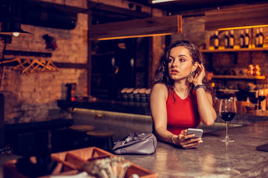 Woman in a red top sitting at a bar with a glass of wine, holding a phone.