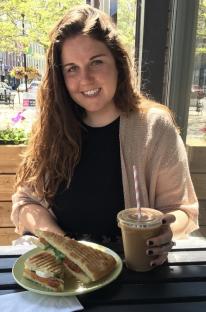 Woman sitting outdoors at a cafe table with a panini and iced coffee.