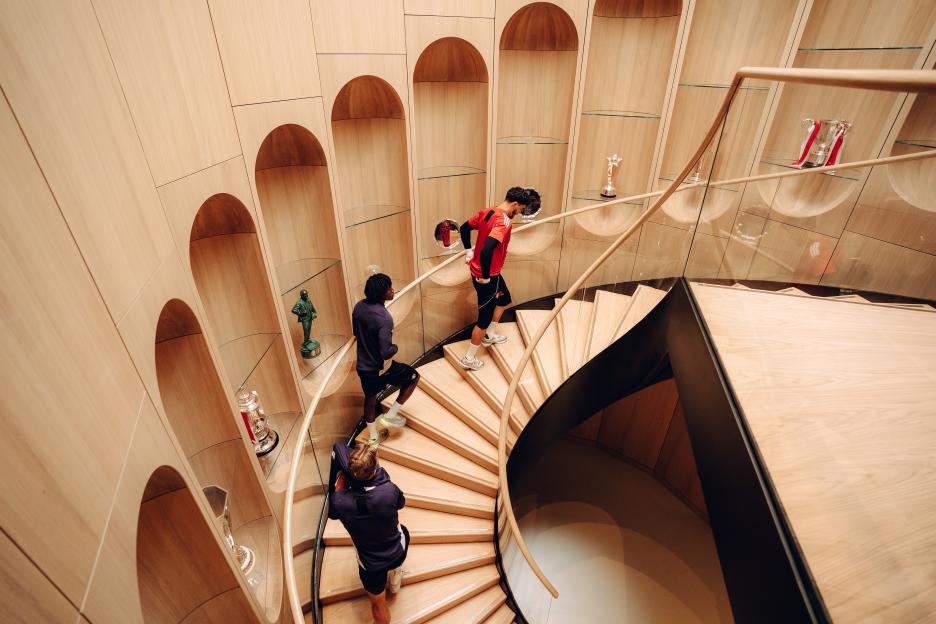Manchester United players ascending a spiral staircase in a building with trophy displays.