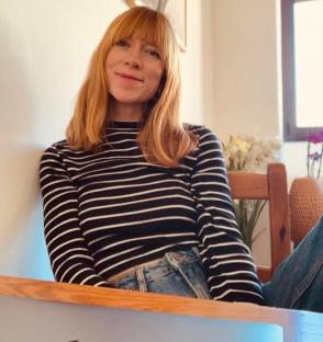 Woman with long, straight, reddish-blonde hair wearing a black and white striped shirt and blue jeans.