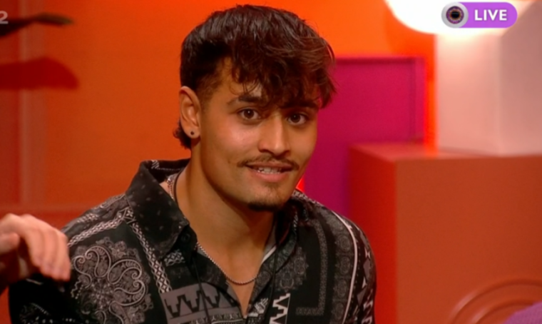 A young man with dark, curly hair and a mustache smiles slightly while wearing a patterned black shirt.