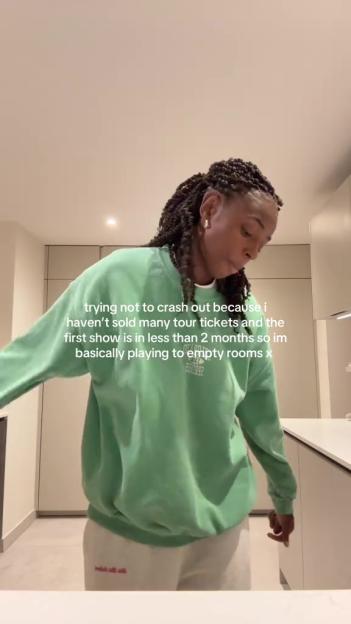 A woman with braided hair wearing a green sweatshirt and white pants, looking down while text on screen discusses not selling many tour tickets.