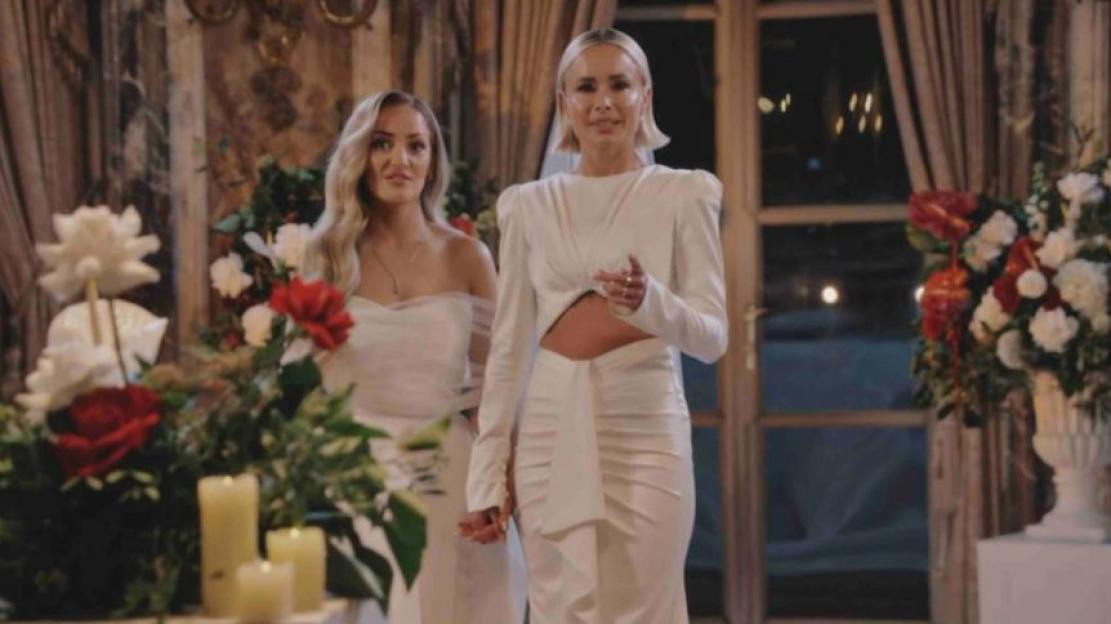 Two brides standing together in white dresses among floral arrangements.