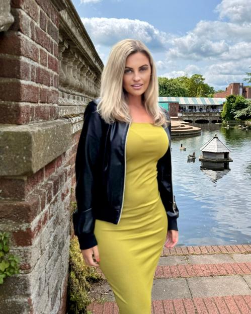 Woman in yellow dress and black jacket by brick wall.
