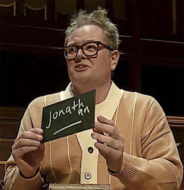 Man wearing glasses and a striped cardigan holding a sign that reads "Jonath RA".