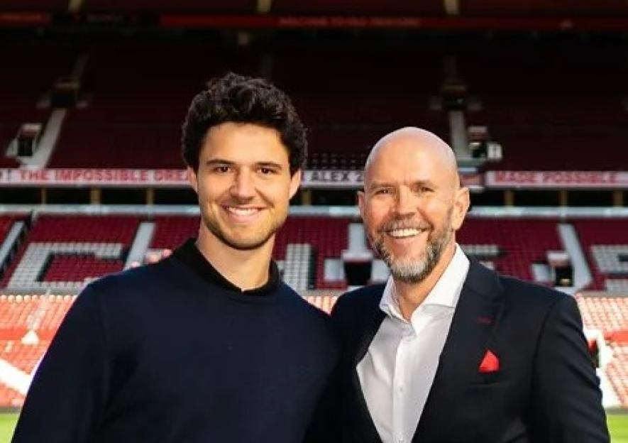 Two men stand on a soccer field in front of Old Trafford stadium.