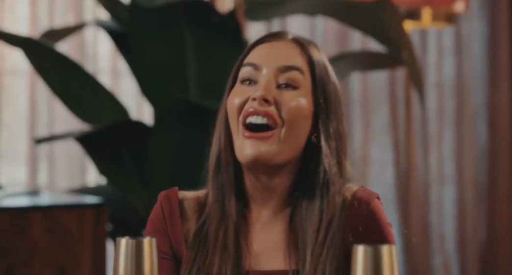 A woman with long brown hair smiles widely, revealing her white teeth, against a blurred background of plants and warm tones.