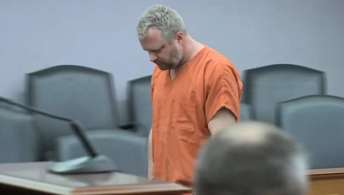 A man in an orange jumpsuit standing in a courtroom, looking down.
