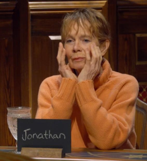 Celia Imrie in an orange sweater, holding her hands to her face, with a place card that reads "Jonathan".