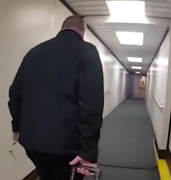 A man in a dark jacket is escorted down an airport hallway.