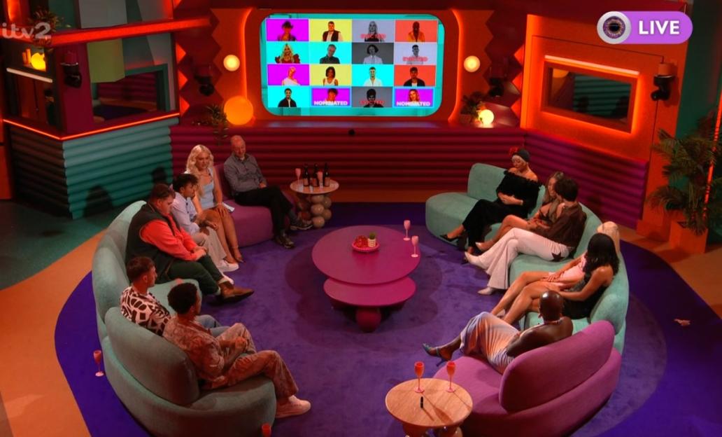 Housemates seated in a living room, watching a screen showing a grid of faces, some labeled "NOMINATED" or "EVICTED."