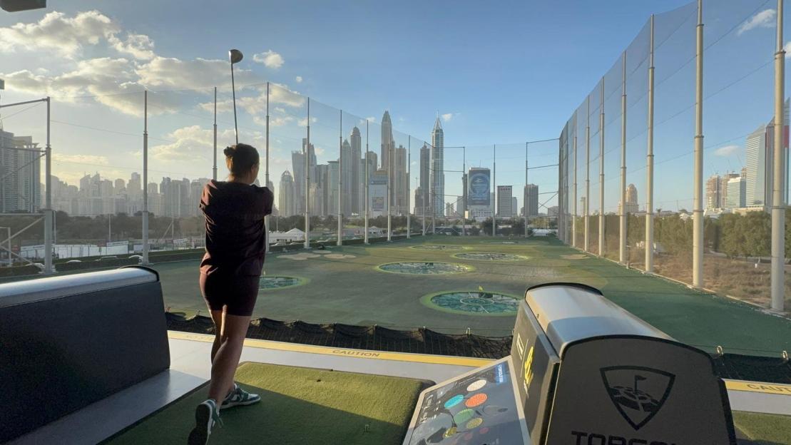 Woman golfing on a driving range with a city skyline in the background.