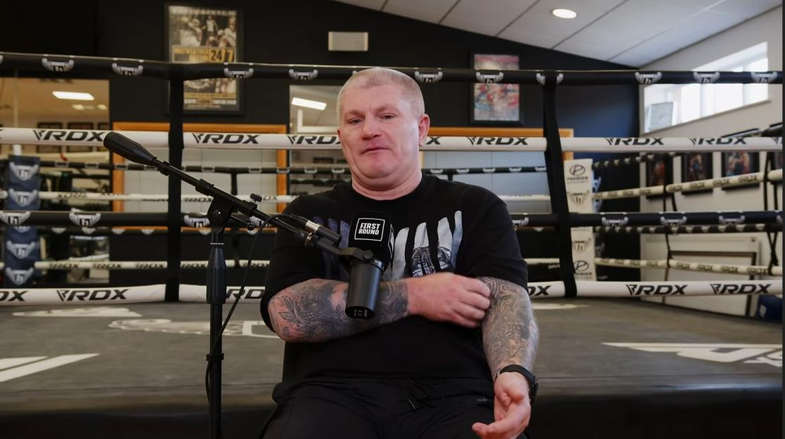 A man with tattoos sits in front of a boxing ring, speaking into a microphone.
