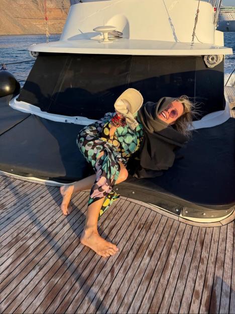 A woman in a colorful dress and sunglasses smiles while lounging on a boat deck.