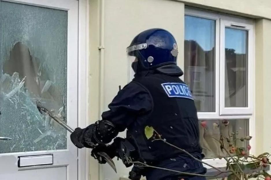 A police officer in riot gear breaking glass in a door.