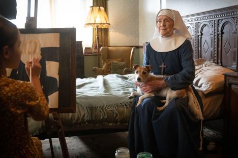 A nun holds a small dog while a young girl paints a portrait.