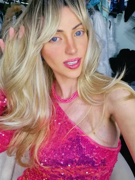 A woman with long blonde hair and bright blue eyes, wearing a pink sequined one-shoulder top and a matching pink necklace.