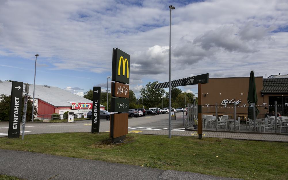 A McDonald's restaurant with a McCafé sign and outdoor seating, with a parking lot and "Einfahrt" (Entrance) sign to the left.