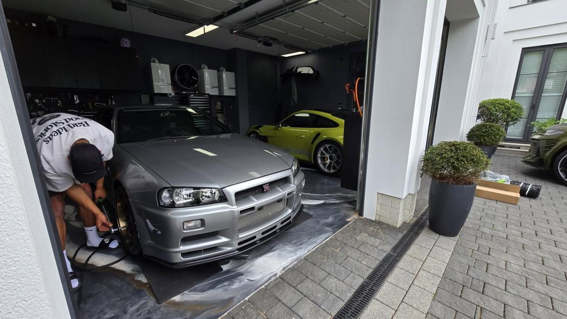 A person cleaning a silver Nissan Skyline R34 inside a garage with a yellow Porsche 911 GT3 RS in the background.