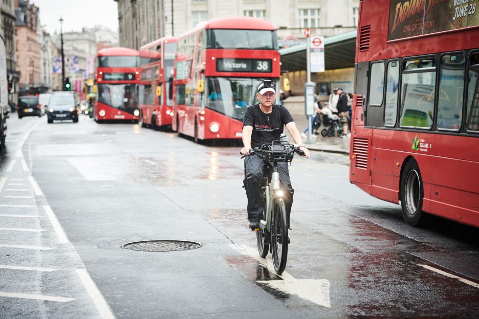 Person cycling in London rain, passing red double-decker buses.