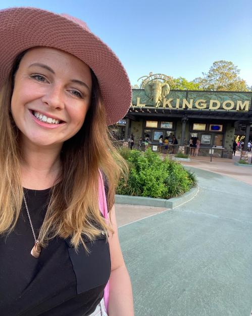 A woman in a pink hat smiles in a selfie in front of the Disney Animal Kingdom entrance.