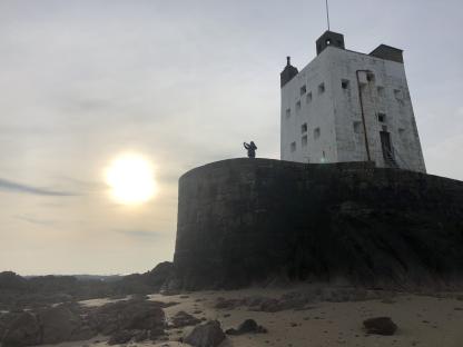 Person taking a picture of a white tower on a rocky coast at sunset.