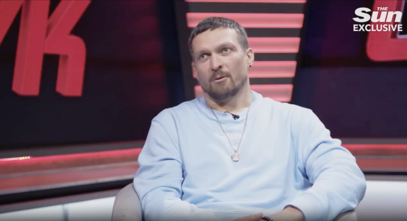 Screenshot of a man in a light blue sweatshirt sitting in a chair during a television interview.