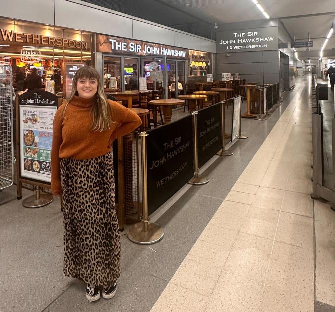 A woman stands in front of "The Sir John Hawkshaw" Wetherspoon pub, smiling.