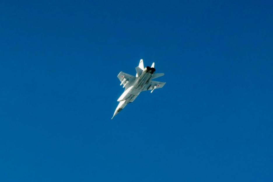 A white Russian MiG-31 fighter jet with red stars on its wings flying in a clear blue sky.