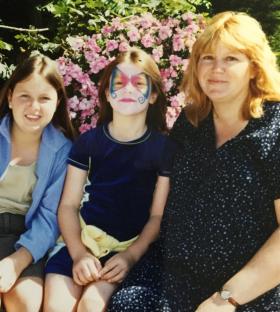 Photo of three women, two young girls and an older woman, sitting together in front of pink flowers. One girl has butterfly face paint.
