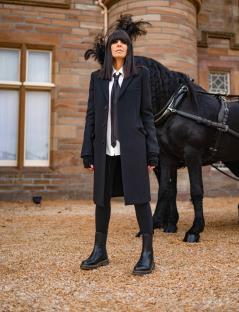 Claudia Winkleman standing in front of a black horse and carriage.