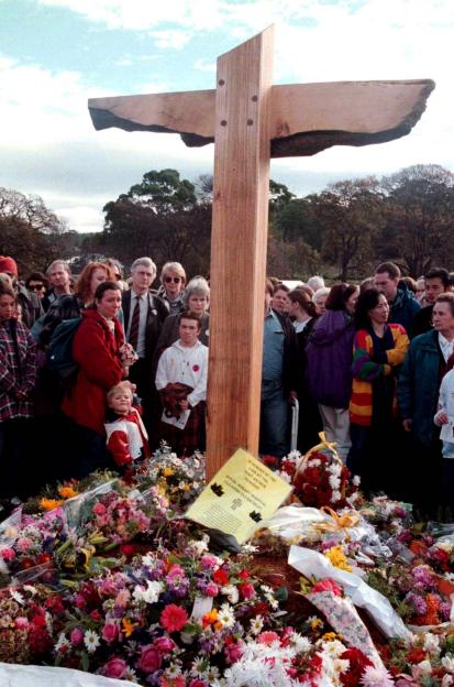 Mourners attend a memorial service at Port Arthur, Tasmania, featuring a wooden cross and flowers.