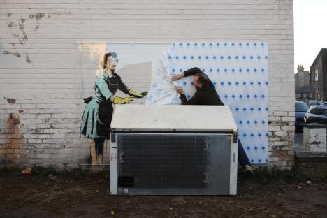 A worker covering a Banksy artwork depicting a woman in a 1950s-style dress with a protective screen.