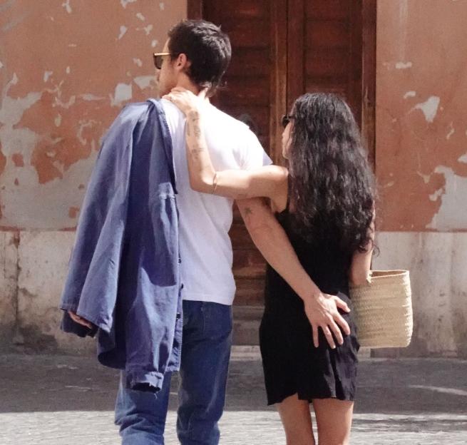Harry Styles and Zoe Kravitz walking away, with Styles in a white t-shirt and blue jacket over his shoulder and Kravitz in a black dress with her hand on Styles' shoulder and his hand on her lower back.