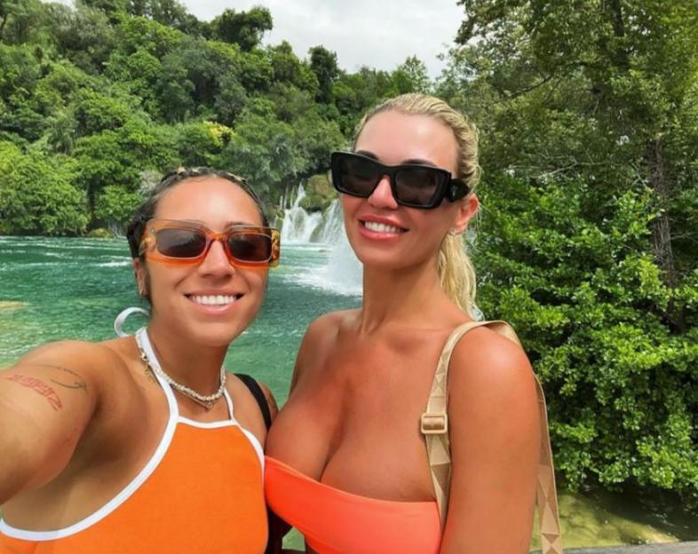 Two women wearing sunglasses in orange tops in front of a waterfall.