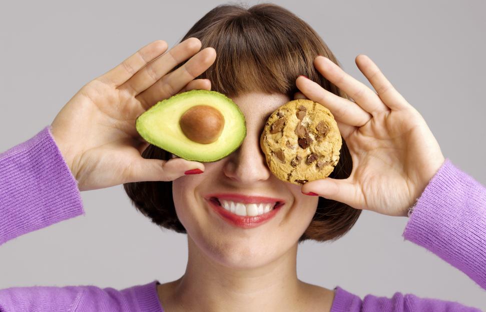 Woman holding an avocado slice over one eye and a chocolate chip cookie over the other, smiling.