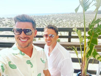 Two men smiling, wearing sunglasses, on a beach patio.
