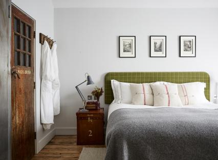 Hotel bedroom with a green headboard, grey blanket, and antique luggage as a nightstand.