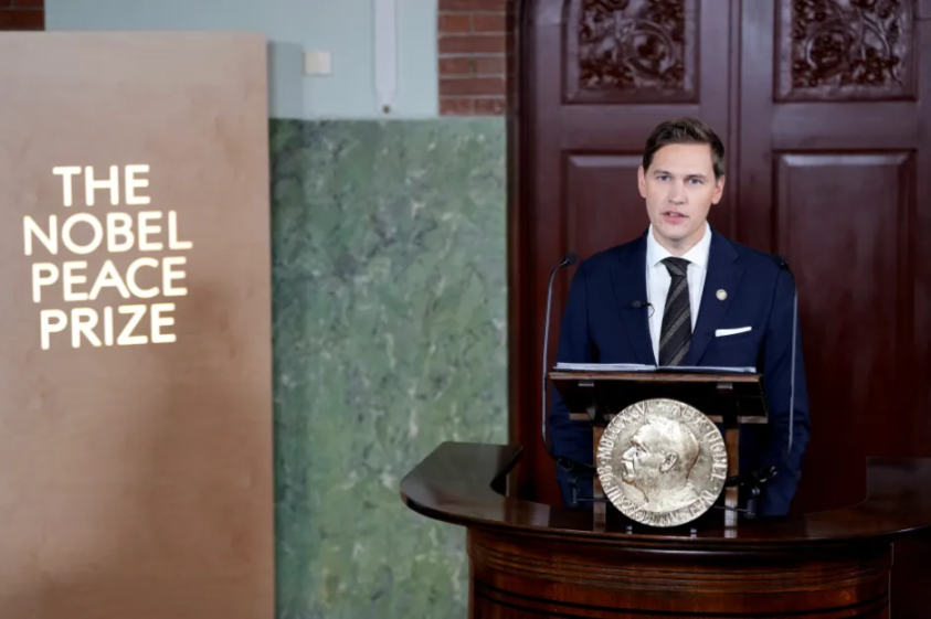 A man in a suit speaking at a podium with "The Nobel Peace Prize" sign in the background.