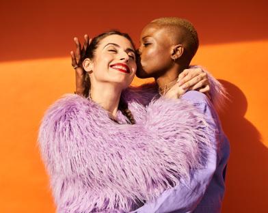 Two women embracing and kissing in front of an orange background.