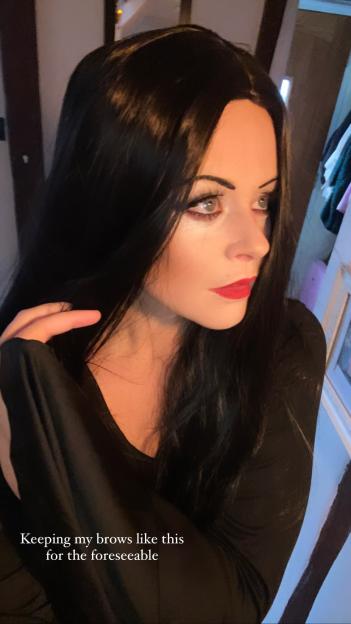 Emily Atack dressed as Morticia Addams, with long black hair, red lipstick, and dark eye makeup.