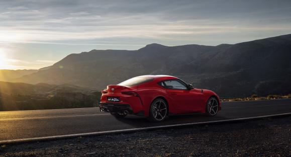 Red Toyota GR MKV Supra driving on a mountain road at sunset.