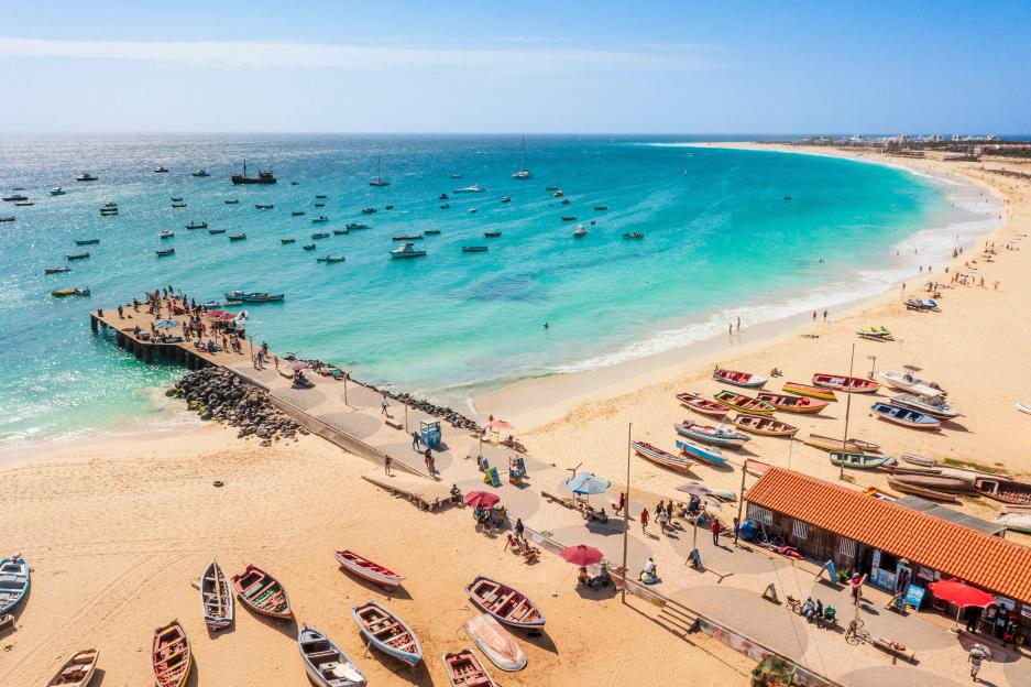 Aerial view of Santa Maria, Cape Verde, showing a pier with people, many boats in turquoise water, and a long sandy beach with more boats and people.
